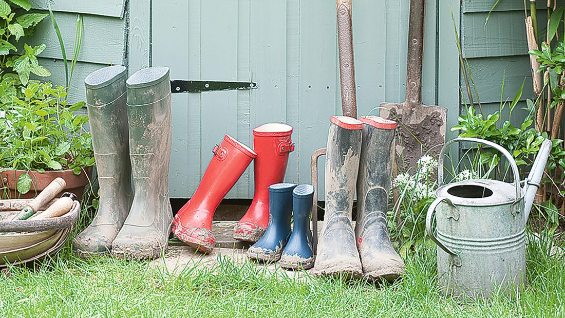 Gardening tools and equipment next to a shed, including gumboots, a shovel, a pitchfork and a watering can.