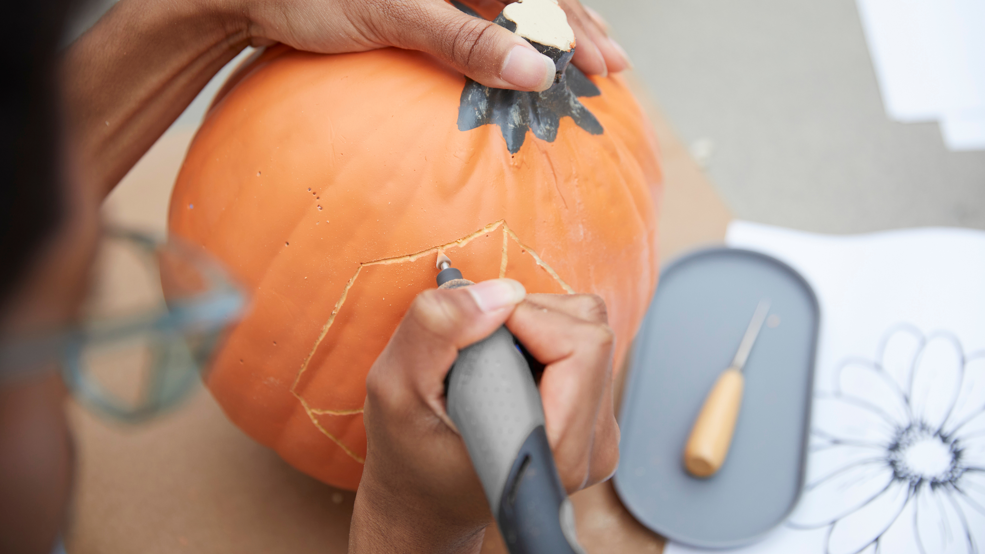 Hand gripping the carving tool and carving designs into a pumpkin.