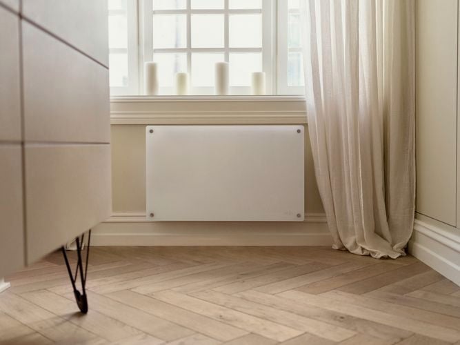 Glass panel heater mounted on wall space between skirting board and a window.