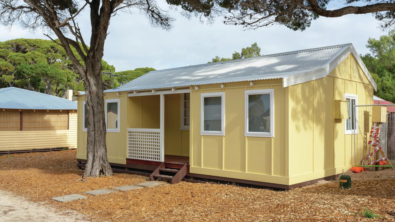 A yellow cabin built on Rottnest island with a white lattice fenced porch and a tree