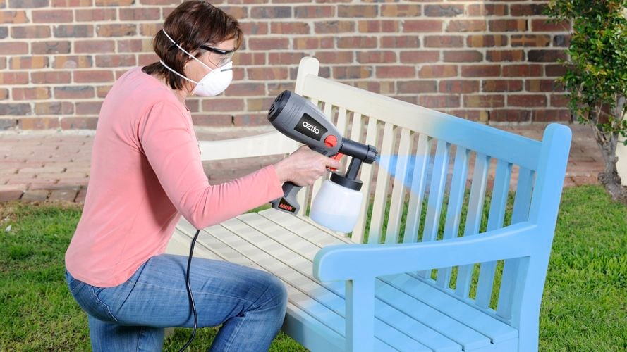 Spray painting wooden outdoor bench a cream shade with an Ozito paint sprayer. Woman is wearing mask and goggles for use.