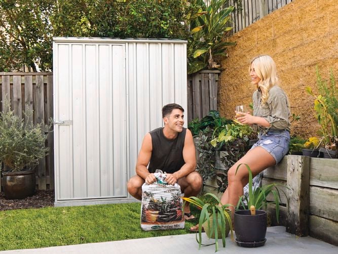 A man crouching holding potting mix and a woman holding at the front of a shed outside.