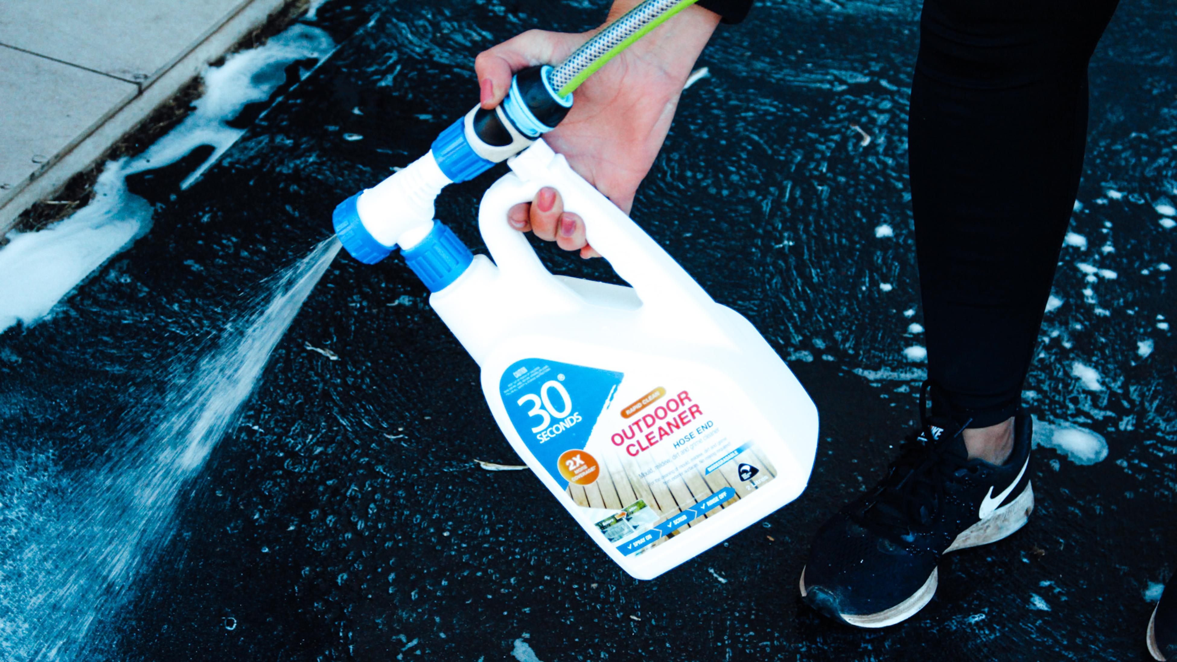 30 seconds outdoor cleaner bottle in a user's hand, bottle is attached to a hose with liquid being sprayed out onto the concrete.