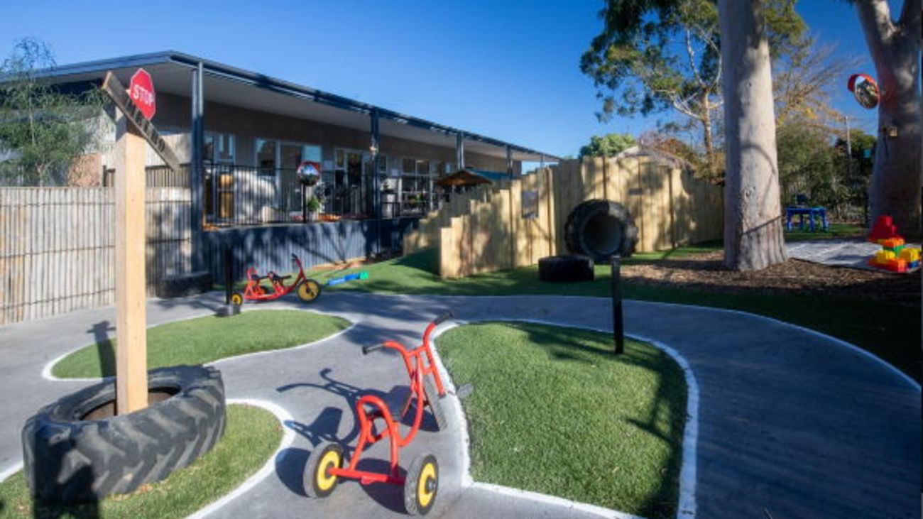 The outdoor playground of a kindergarten, with tricycle, concrete track, tanbark area and big tree, in front of a large building.