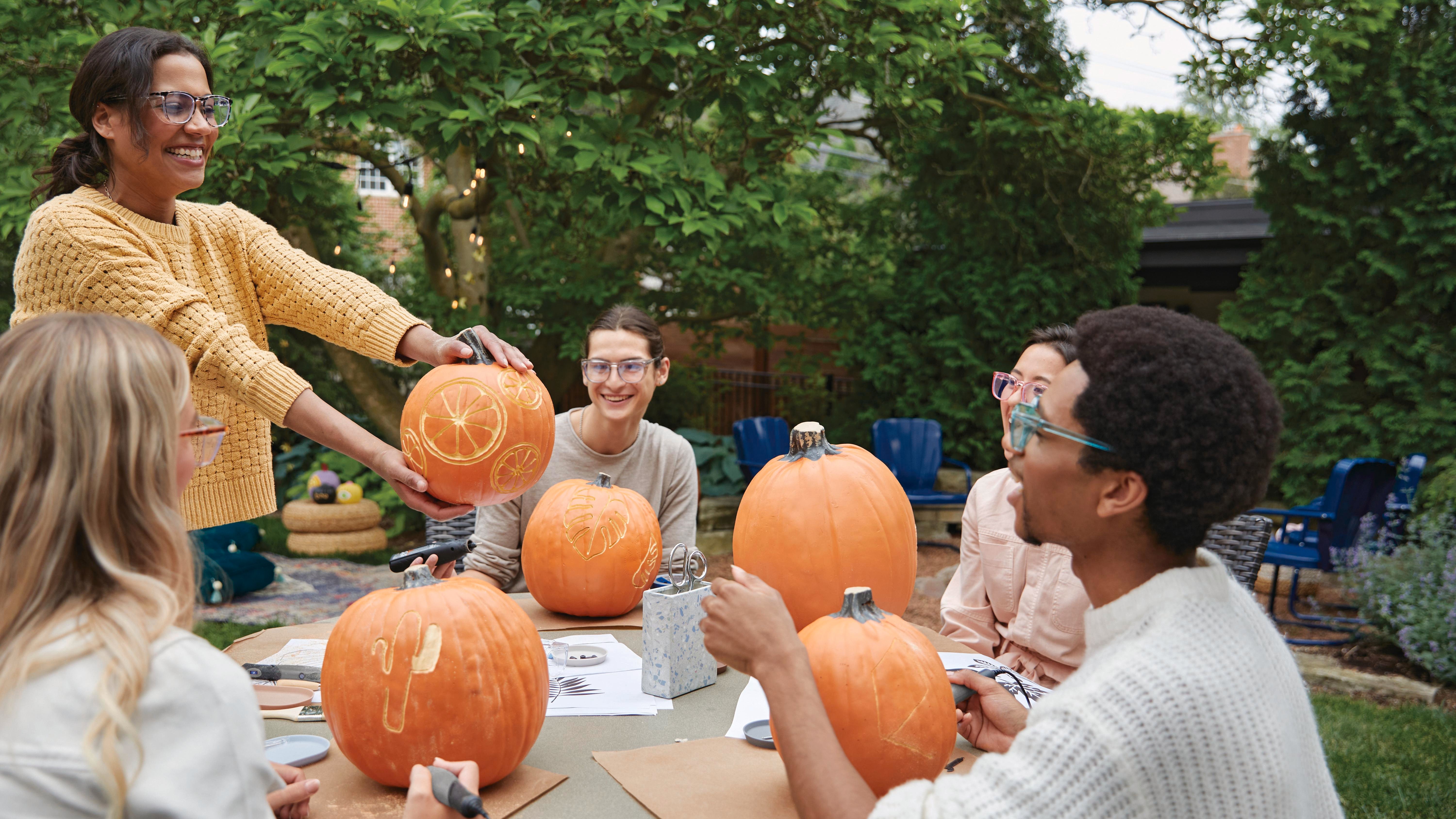 Group of people sitting outdoors at a wooden table, carving pumpkins.