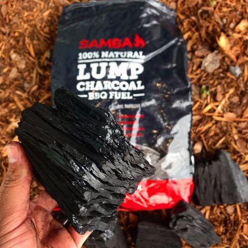 Person holding barbecue charcoal in front of a bag of Samba '100% Natural Lump Charcoal BBQ Fuel'