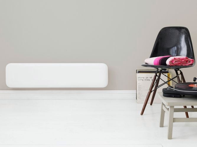White slim panel heater mounted on a wall with a black chair to the side.