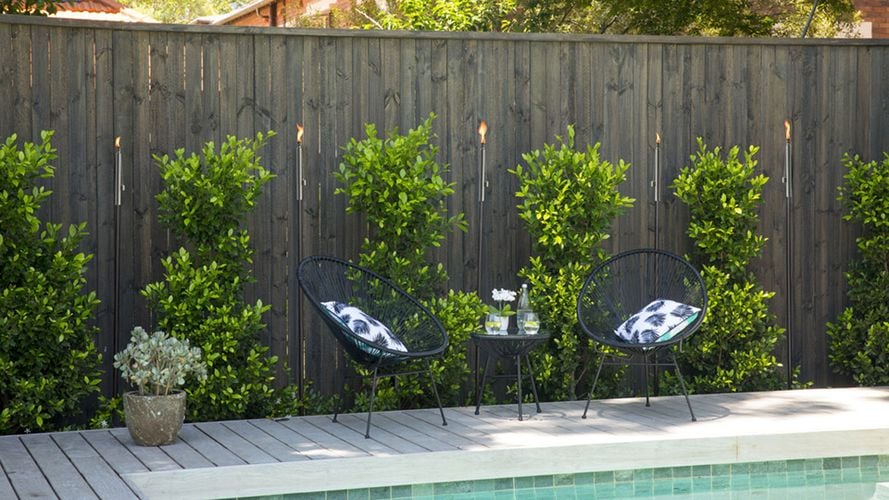 Flaming torches interspersed with trees against a wooden fence behind an outdoor lounge setting