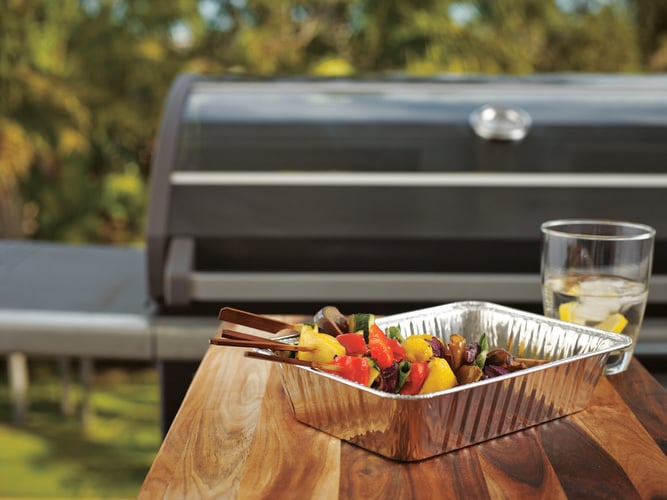 Foil tray containing meat and vegetables in front of barbecue